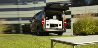 Hnědá dodávka s otevřenými zadními dveřmi a skládacím stolem před ní.