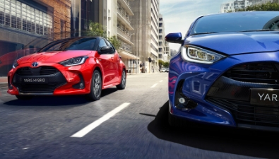Dvě červené a modré Toyoty Yaris Hybrid jedoucí po městské ulici.